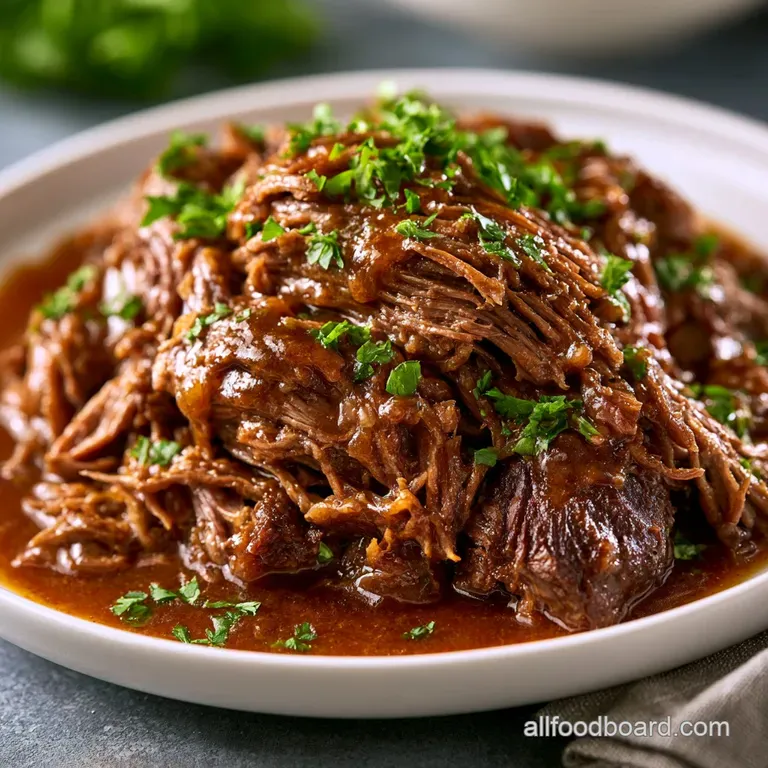 Juicy, slow-cooked beef on a white platter, drizzled with glossy gravy and garnished with sliced green peppers.